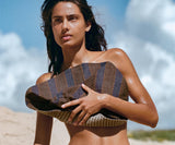 Woman in a striped bikini top and black bottoms holding a clutch on a beach.