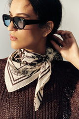 A woman wearing black sunglasses, a brown knit sweater, and a cream bandana scarf with a brown landscape print, shown in a warm side-profile portrait.