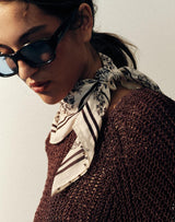 A woman wearing a dark brown open-knit poncho-style sweater, a patterned scarf, and sunglasses, photographed against a plain light background.