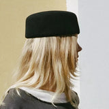 A person shown in side profile wearing a black felt beret-style hat, with long blonde hair and a dark gray sweatshirt, standing against a split beige and white wall.