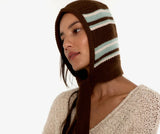 A woman wearing a brown knit bonnet with light blue and white stripes, paired with a chunky cream-colored knit sweater, against a plain white background.