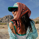 Woman sitting outdoors in a desert-like setting wearing a green cap, striped shirt, and colorful headscarf.