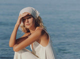 A woman wearing a flowing white maxi dress and matching headscarf sits on rocks by the ocean, shielding her eyes from the sun.