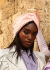 Two models wearing pink and green turban hats with wavy details, posing against a stone wall background.