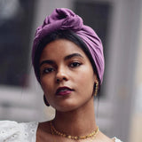Woman wearing a purple headscarf and gold jewelry against a blurred background