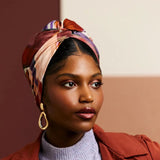 Woman wearing a patterned headscarf, red jacket, and gray sweater against a two-tone wall.