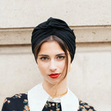 Woman wearing a black turban and floral dress with a white collar against a light-colored wall.
