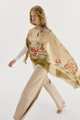 A woman modeling a lightweight beige shawl or poncho with a red and green floral print, worn over a white long-sleeve top and beige wide-leg trousers against a plain white studio background.