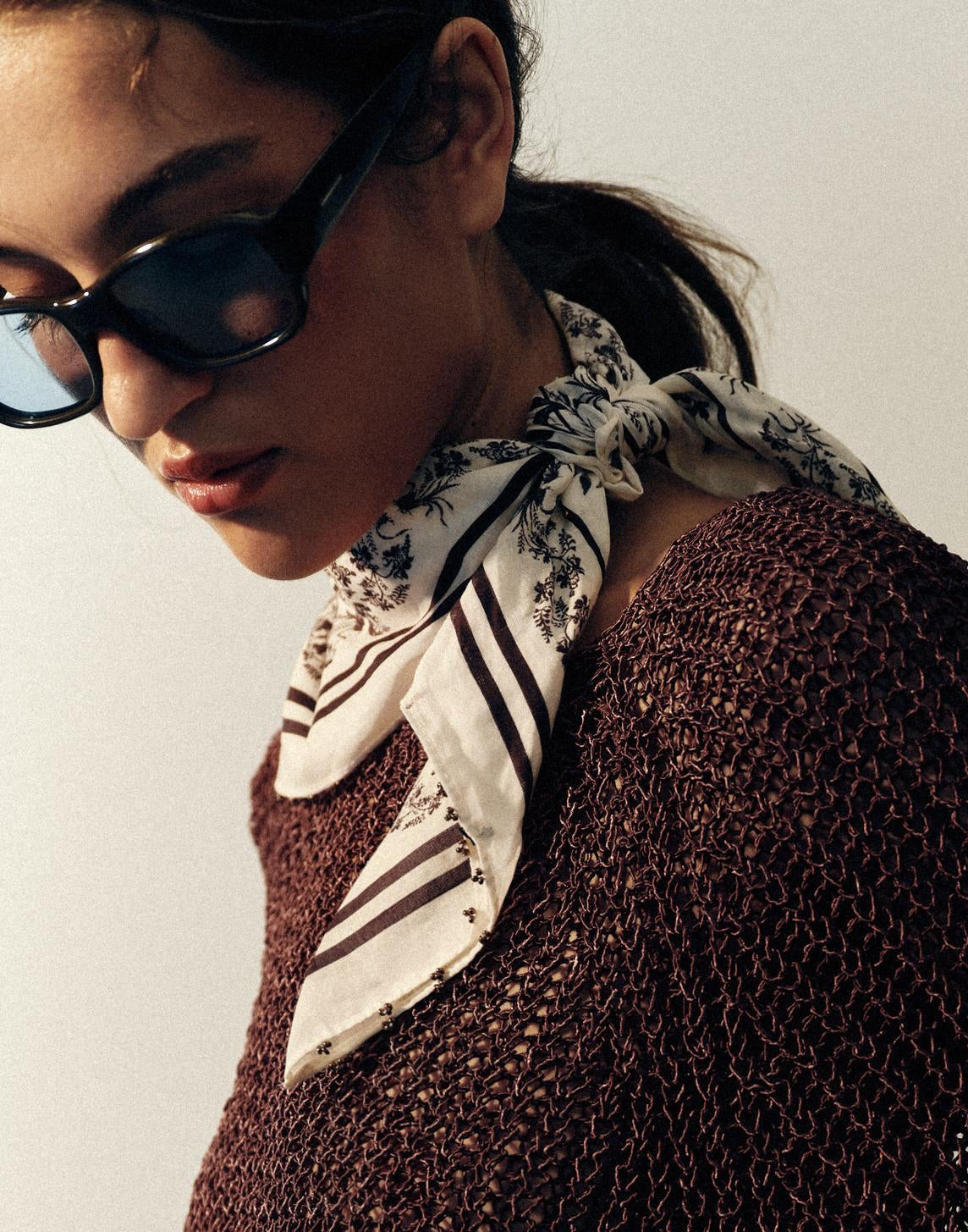 A woman wearing a dark brown open-knit poncho-style sweater, a patterned scarf, and sunglasses, photographed against a plain light background.