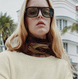 Person wearing a beige sweater and brown pants with sunglasses, standing on a street.