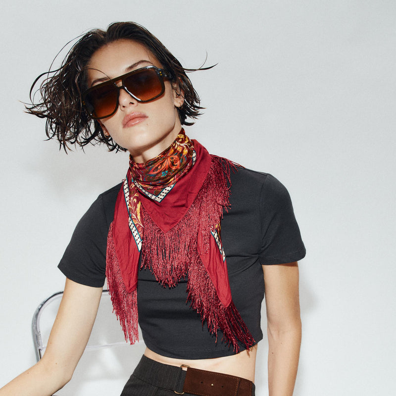 A model wearing a black crop top, black trousers, a brown belt, and a red patterned scarf with fringe, seated on a metal chair against a light gray background.
