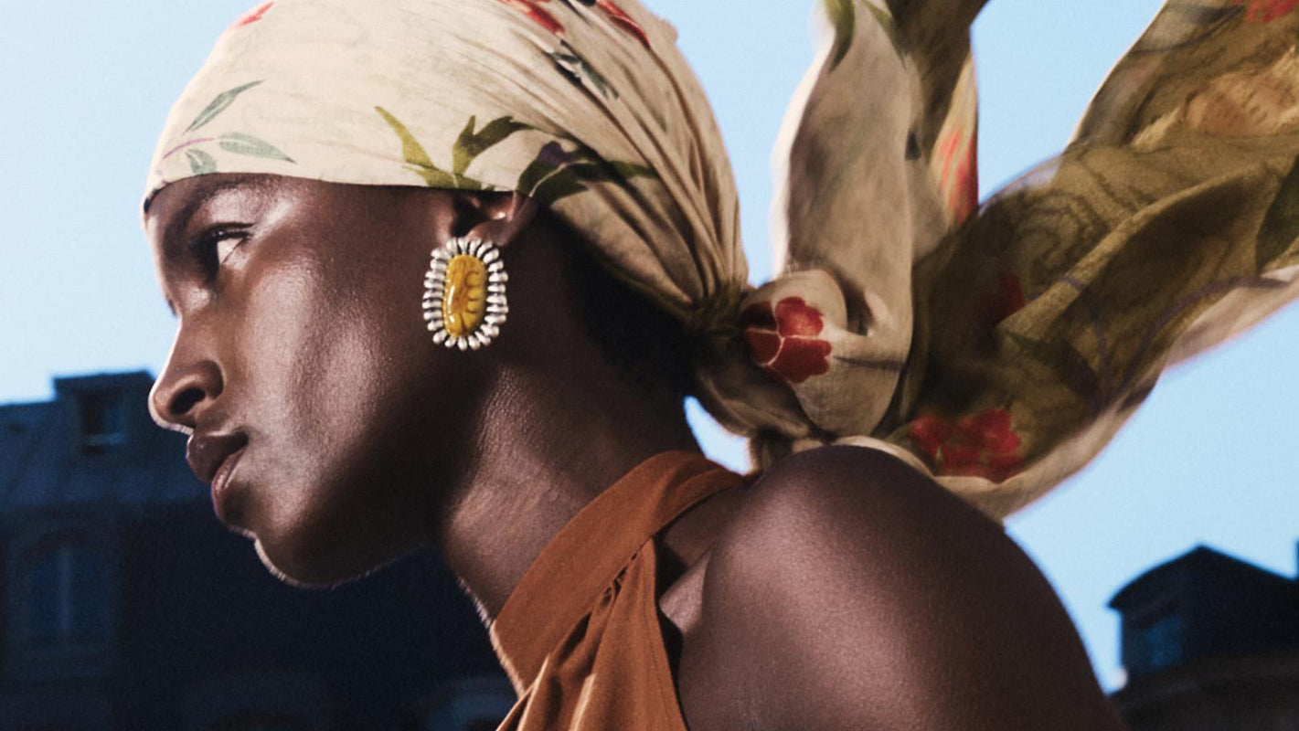 A woman in profile wearing a rust-orange halter top and carrying a large olive-brown shoulder bag, with a beige floral headscarf and gold earrings, set against a blurred city building background.