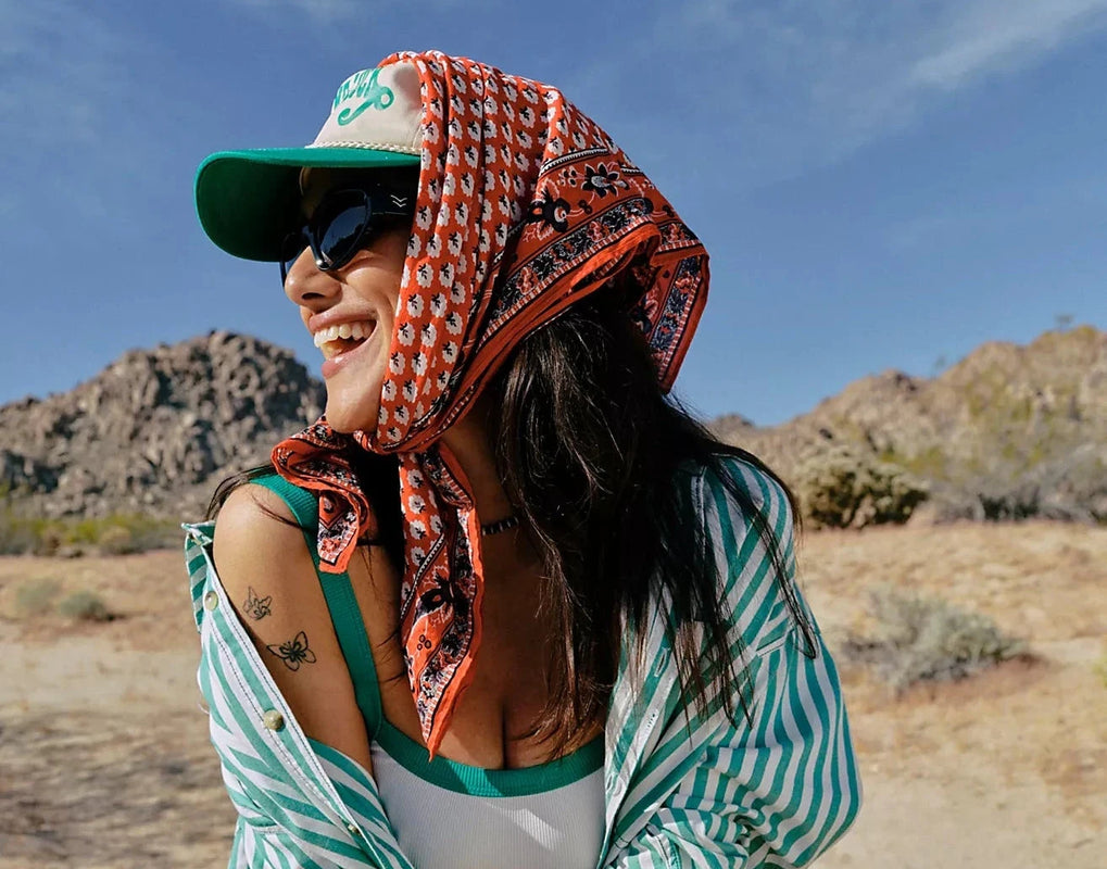 Woman sitting outdoors in a desert-like setting wearing a green cap, striped shirt, and colorful headscarf.