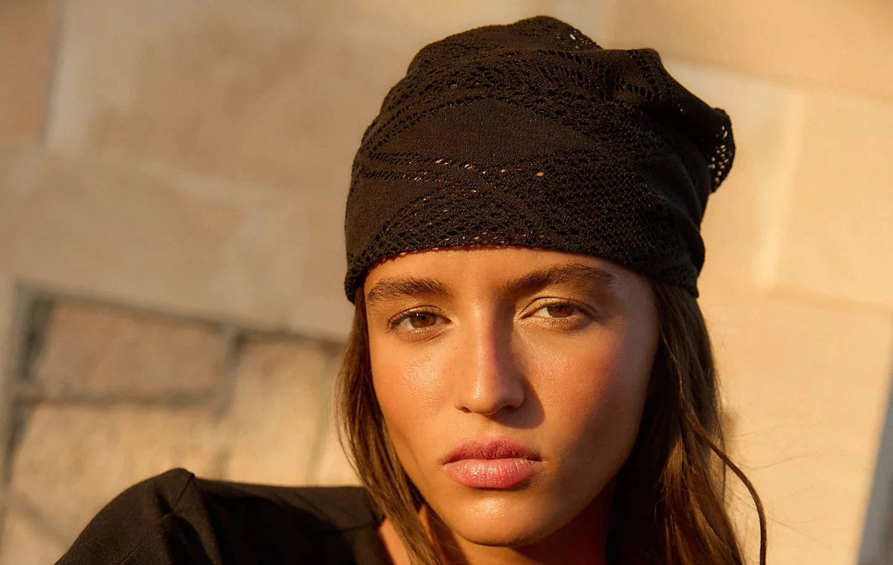 A woman wearing a black knit beanie and a black long-sleeve top, sitting outdoors with her arms crossed. The warm sunlight casts a strong shadow on the background wall.