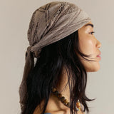 A woman in profile wearing a taupe-beige crocheted headscarf tied under her head, styled with a blue lace-trim camisole and layered necklaces against a neutral beige background.