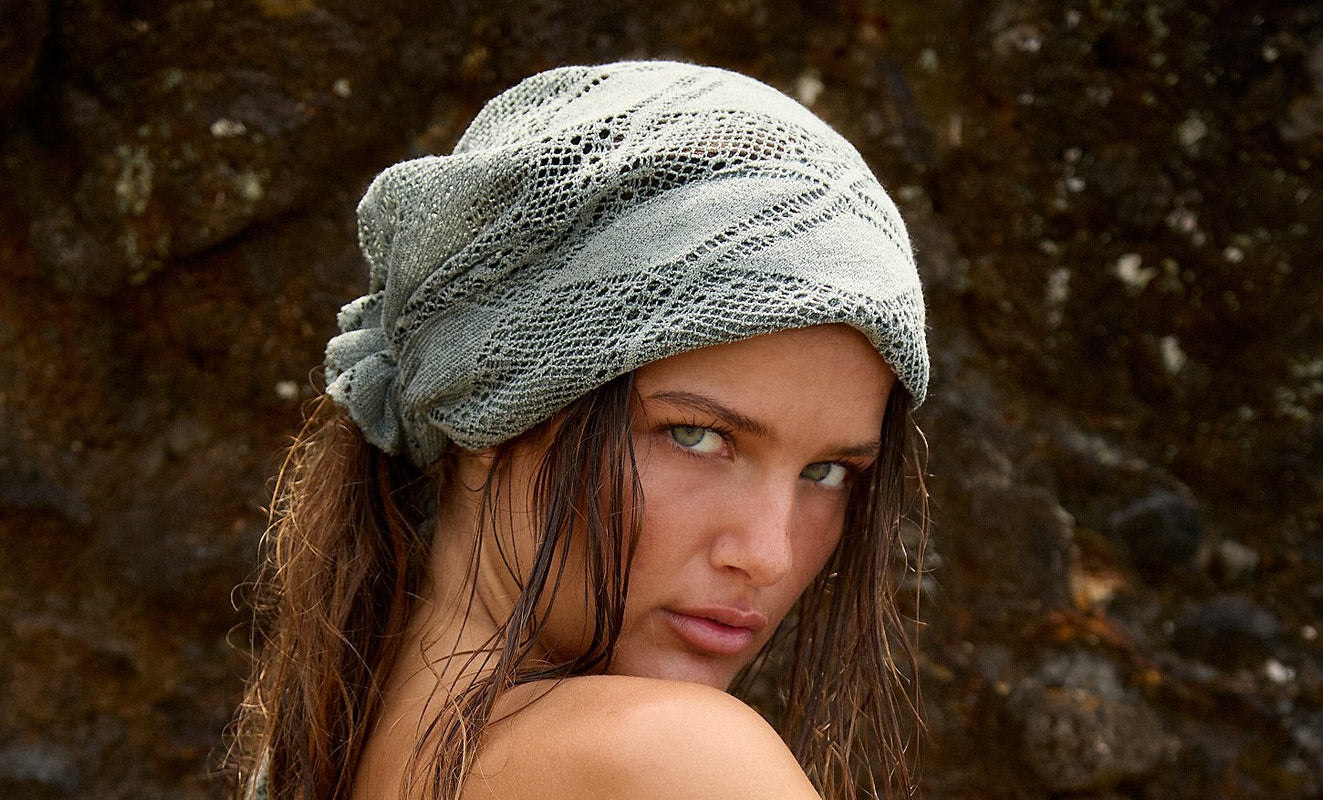A woman wearing a light blue ruffled tube top and a gray knit head wrap, posed against a dark rocky background.