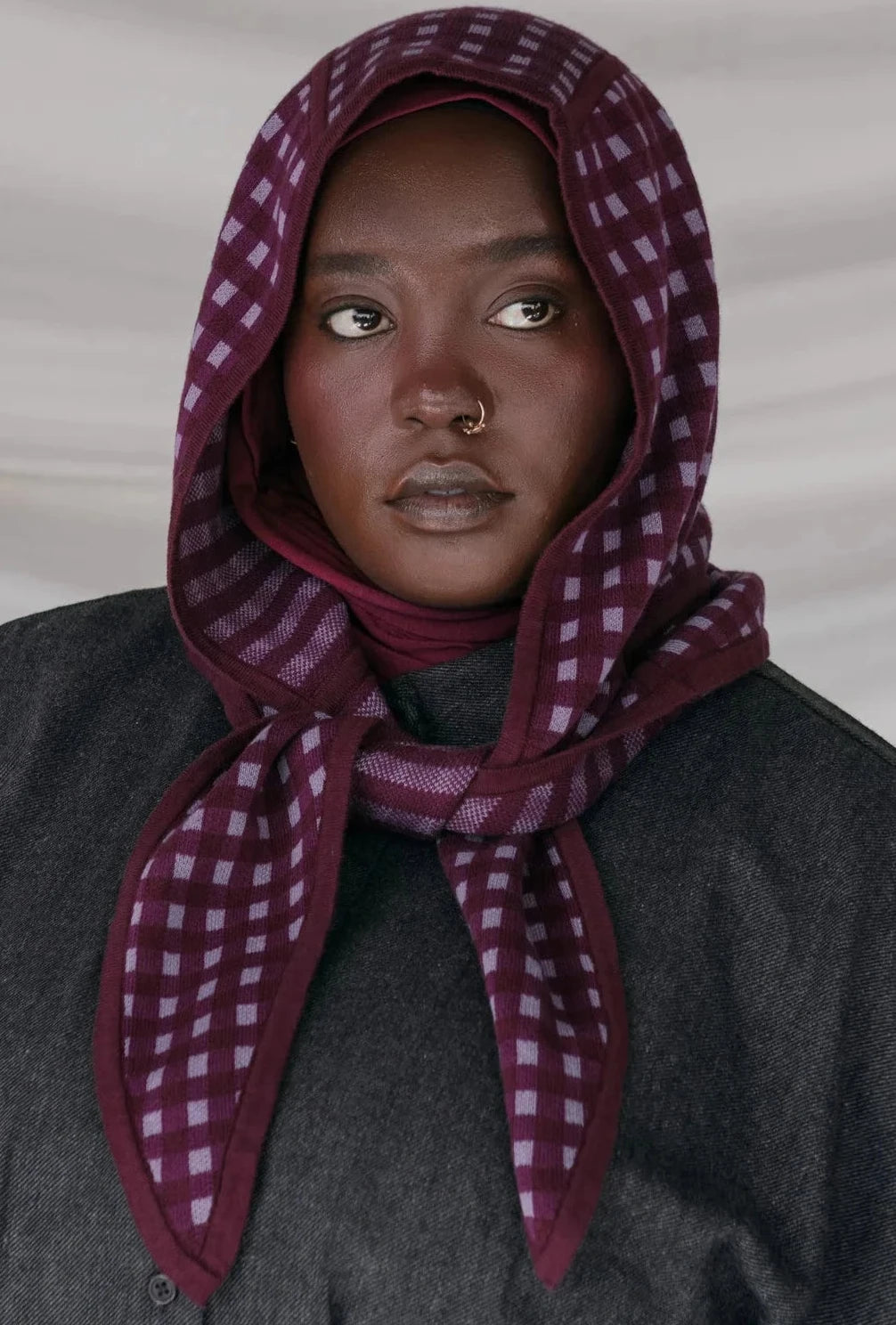 Person wearing a dark gray coat and a purple checkered headscarf against a plain background