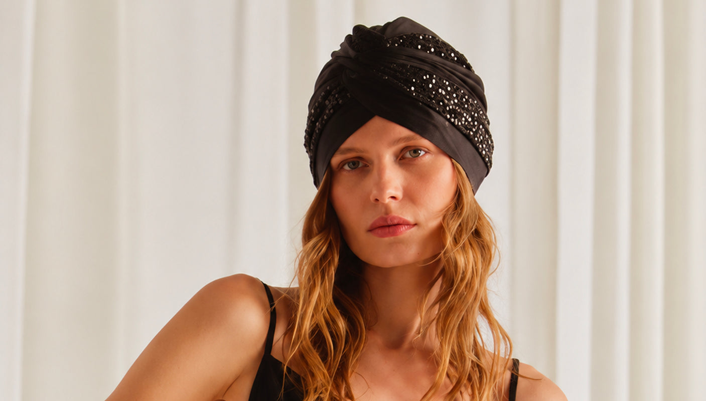 A woman with long wavy light brown hair wearing a black satin turban hat embellished with sparkling rhinestones, set against a soft, off-white curtain background.