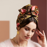 Woman wearing a patterned headscarf with flowers, gold jewelry, and a pink sweater.