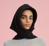 Woman wearing a black hooded top and light gray long-sleeve shirt against a pink background